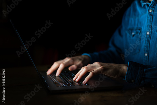Man working on laptop computer in the dark. Working at night with computer screen light. Blue shirt young business man typing on his computer