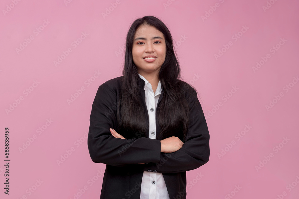 A young southeast asian female employee in a black jacket with arms crossed. Isolated on a light pink background. A young southeast asian female employee in a black jacket with arms crossed. Isolated on a light pink background.