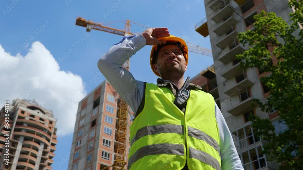 Happy male foreman in vest putting on yellow hardhat while posing at construction site with new building on the background. Architecture concept. Real time