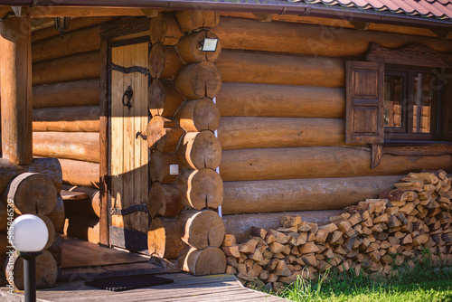 A neat large woodpile against the background of a wooden house made of logs with a small window. Sunny spring or summer day