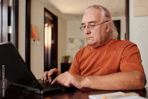 Senior man working on laptop at home, answering e-mails and filling documents