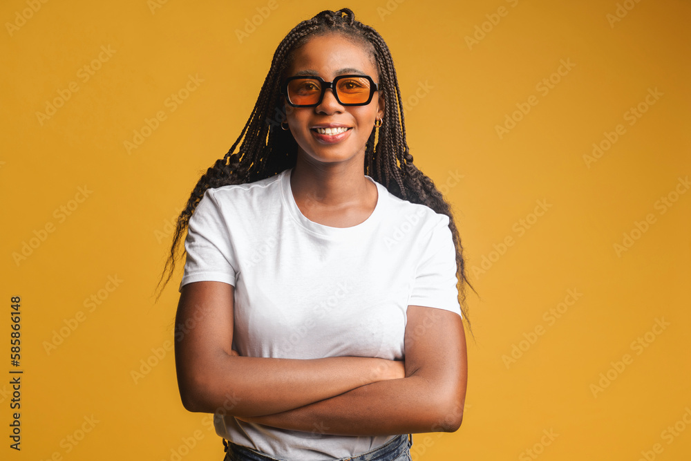 Charming young brunette woman stands with arms crossed and looks at the camera, beautiful positive african american lady smiles isolated on yellow background