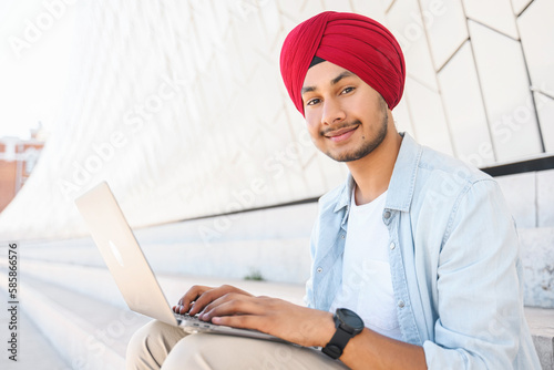 Optimistic Indian handsome guy in red turban using laptop outdoors. Ethnic hindu freelancer man sitting on the steps in city landscape and looking at the camera, smiling friendly photo