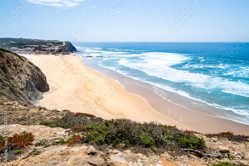 A panoramic high angle view from a steep cliff over Praia de Monte Clérigo beach, small village Monte Clérigo and the horizon over the Atlantic Ocean on a sunny day in May with soft light from behind.