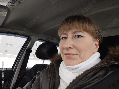 Portrait of adult cute funny blonde woman in warm clothes taking a selfie inside the car and driving it. The concept of a pleasant trip in spring, autumn, winter
