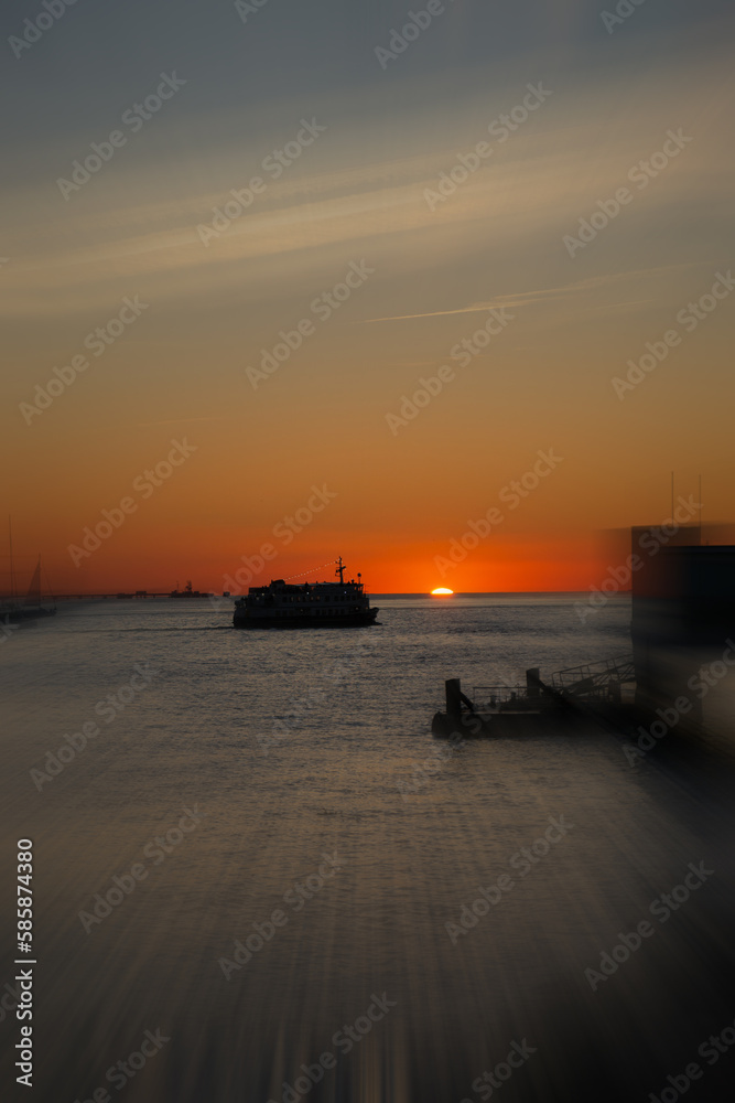 sunset with a silhouetted boat sailing just out of port along its journey against a vivid colorful sunset an orange and yellow color filled sky with motion blur.