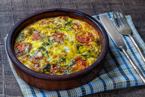 Ceramic bowl with vegetable frittata, simple vegetarian food. Frittata with egg, tomato, pepper, onion, green dill and cheese on wooden table, closeup. Italian egg omelette