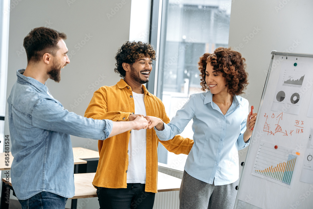 Obraz premium Group of a cheerful multiracial colleagues, stand in a office near a marker white board, analyze the results of work, financial profit, rejoice in success, all together touch with their fists, smile