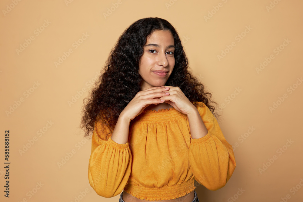 Studio portrait of pretty shy young adorable sweet girl with long wavy hair in yellow clothes standing against brown studio background with folded palms under chin, looking at camera with cute smile Studio portrait of pretty shy young adorable sweet girl with long wavy hair in yellow clothes standing against brown studio background with folded palms under chin, looking at camera with cute smile