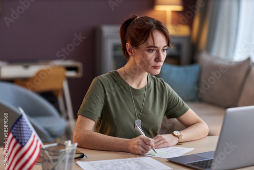 Portrait of young military woman looking at laptop screen and taking notes in online meeting