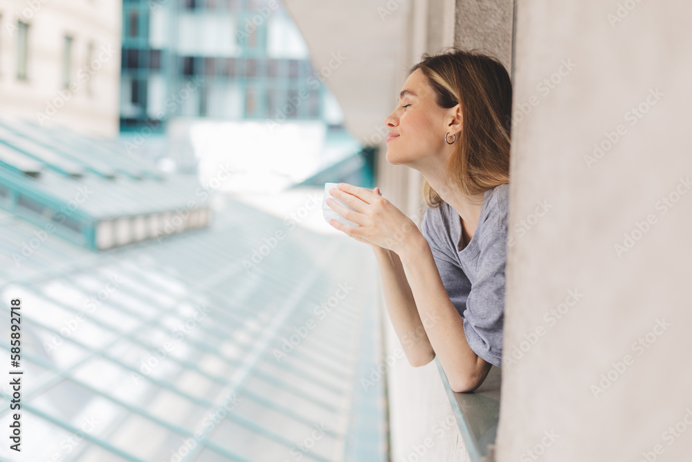 Fototapeta premium Young woman looking out the window and holding mug at home or hotel room. She drinking coffee or tea after wake up in the morning. Woman look enjoy and happy, positive mood, smell of morning coffee