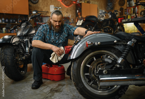 Bearded mature man biker cleaning motorcycle in garage workstation