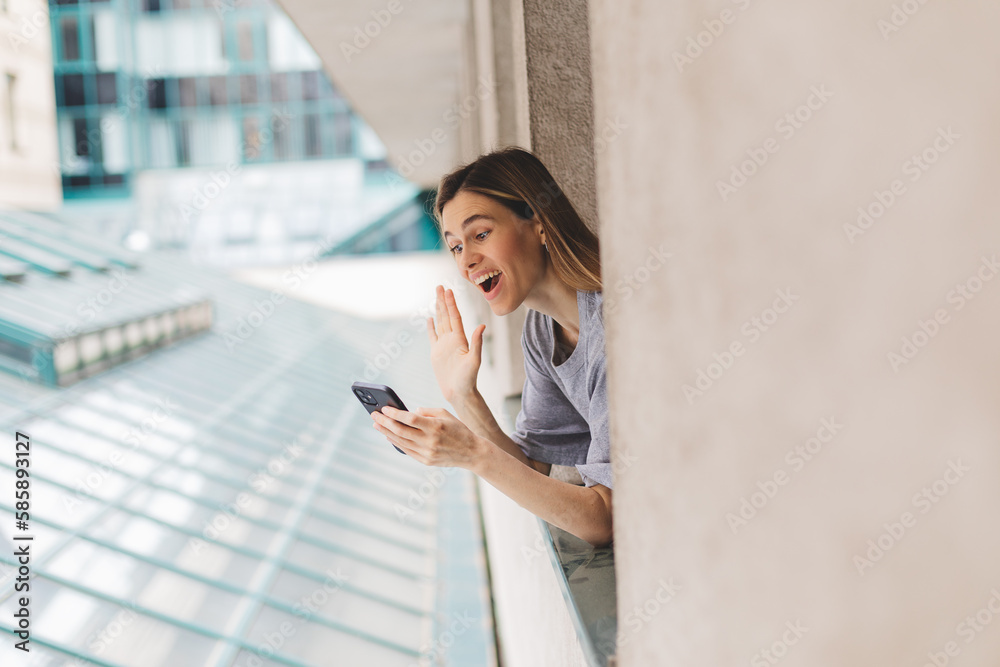 Fototapeta premium Young woman looking out the window and holding smart phone at home or hotel room. Woman using mobile phone chatting, video call or message, zoom in the morning. Woman look enjoy and happy, hi gesture.