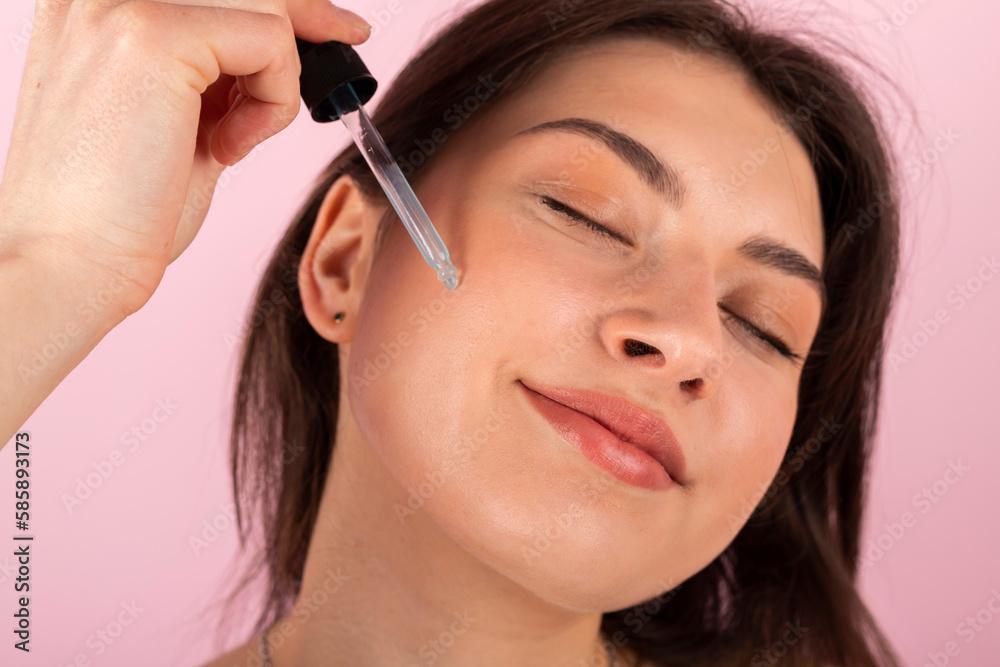 An attractive teenage girl wrapped only in a white towel is happily smiling and using a new cosmetic product on her face with a dropper. isolated on a pink background. Close-up. An attractive teenage girl wrapped only in a white towel is happily smiling and using a new cosmetic product on her face with a dropper. isolated on a pink background. Close-up.