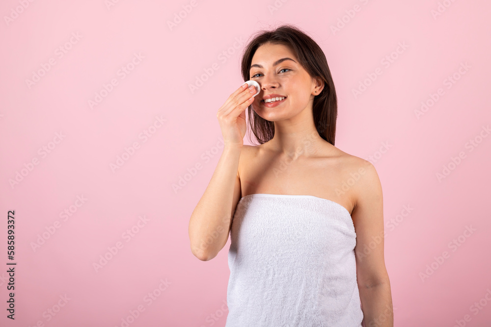 An attractive teenage girl wrapped only in a white towel is happily smiling and using a cotton round or pad. isolated on a pink background. An attractive teenage girl wrapped only in a white towel is happily smiling and using a cotton round or pad. isolated on a pink background.