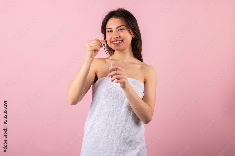 An attractive teenage girl wrapped only in a white towel is happily smiling and holding a new cosmetic product in a small bottle with a dropper. isolated on a pink background. An attractive teenage girl wrapped only in a white towel is happily smiling and holding a new cosmetic product in a small bottle with a dropper. isolated on a pink background.