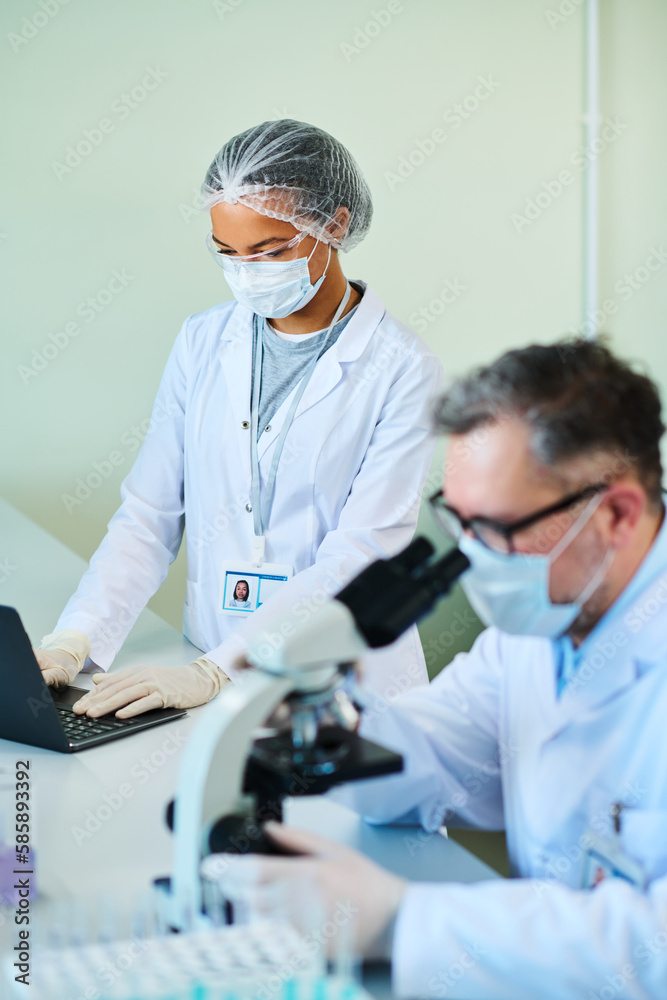 Focus on young female assistant of experienced scientist typing on laptop keyboard while standing by desk next to mature male virologist Focus on young female assistant of experienced scientist typing on laptop keyboard while standing by desk next to mature male virologist