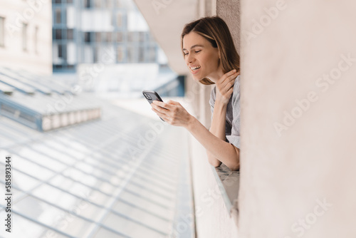 Young woman looking out the window and holding smart phone at home or hotel room. Woman using mobile phone chatting, scrolling, read news in the morning. Woman look enjoy and happy, positive mood.
