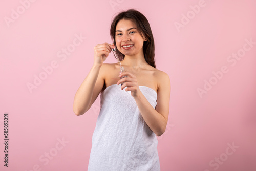 An attractive teenage girl wrapped only in a white towel is happily smiling and holding a new cosmetic product in a small bottle with a dropper. isolated on a pink background.