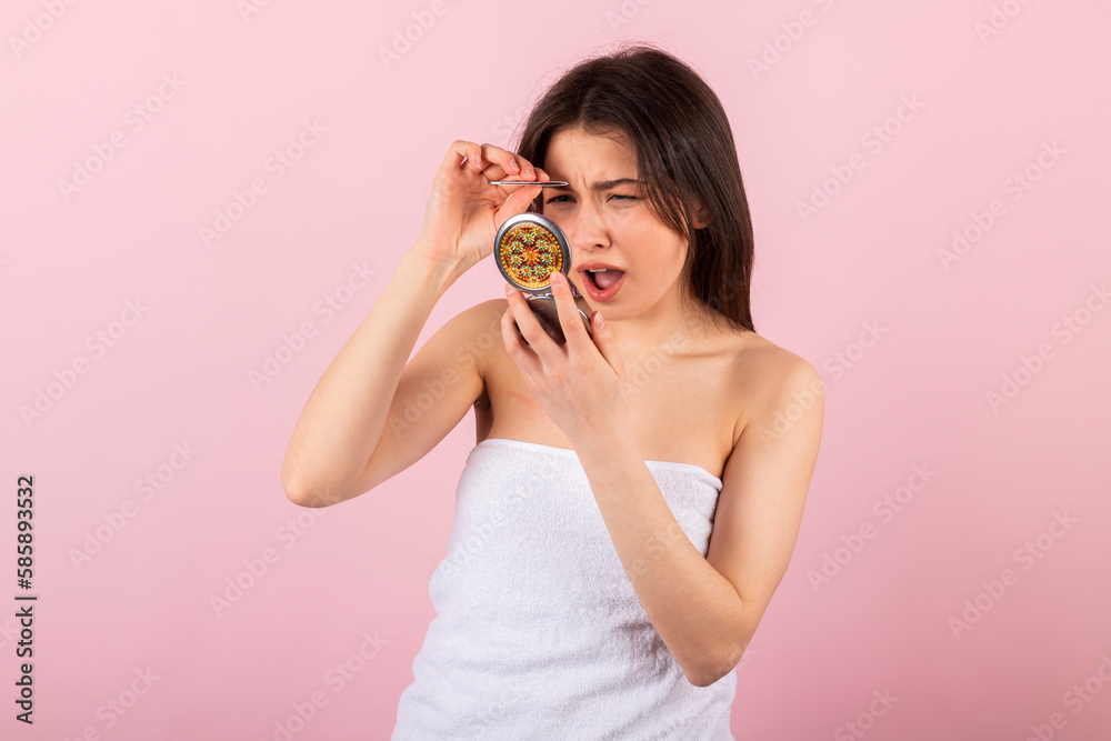 attractive teenage girl wrapped only in a white towel is looking at herself in a small portable mirror and plucking her eyebrows with tweezers. isolated on a pink background. attractive teenage girl wrapped only in a white towel is looking at herself in a small portable mirror and plucking her eyebrows with tweezers. isolated on a pink background.