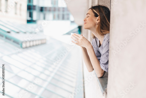 Young woman looking out the window and holding mug at home or hotel room. She drinking coffee or tea after wake up in the morning. Woman look enjoy and happy, positive mood, smell of morning coffee.