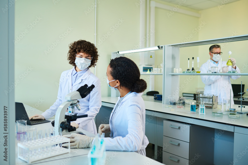 Two intercultural female virologists in lab coats and protective masks having discussion of characteristics of new virus in laboratory Two intercultural female virologists in lab coats and protective masks having discussion of characteristics of new virus in laboratory