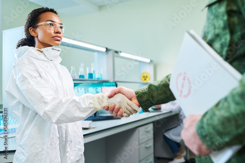 Young serious woman in white hazmat suit shaking hand of military man in camouflage uniform holding folder with secret documents