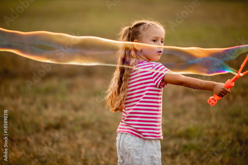 cute little girl plays in the summer outdoors, blowing a big multi-colored soap bubble. the child has fun on vacation, children's leisure, fun, games in nature, holiday, children's birthday.