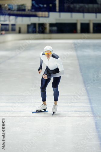 Young professional speed skater in sports uniform having training on ice rink while bending slightly forwards and sliding along arena