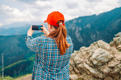 Young woman is standing on the summit of a mountain, taking a photo with a camera. Woman is positioned at the very top of the mountain, with a breathtaking scenic view behind her. 