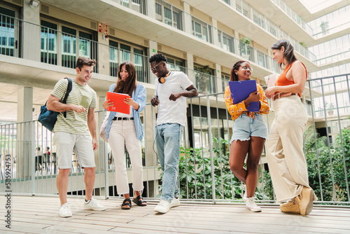 Multiracial group of adolescent students talking together about the exam and homeworks. Happy classmate teenagers smiling taking a breack standing at university campus holding notebooks and backpacks photo