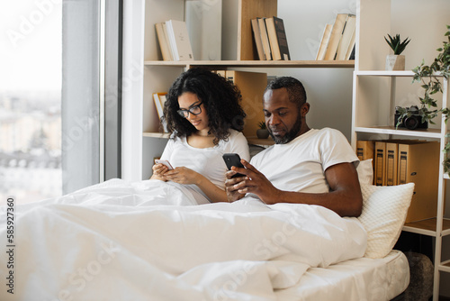 Serious multicultural husband and wife surfing internet using smartphones while sitting under cozy duvet in bedroom of modern flat. Busy spouses networking through social media via digital devices.