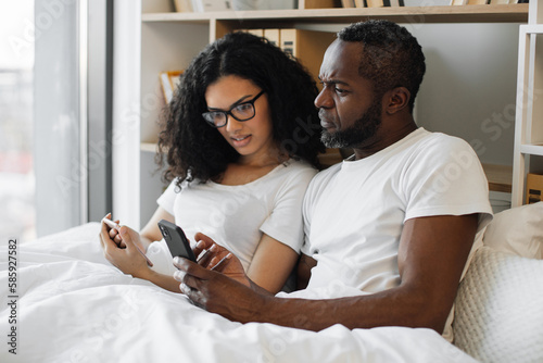 Serene multiracial family of two using mobile phones while lazing in soft bed during daytime at home. Confident adult man and bespectacled woman reading online article paying attention to details.