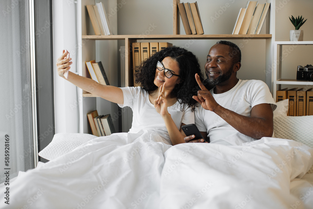 Smiling intercultural woman in glasses taking selfie of her and her husband showing peace gesture while relaxing in bed at home. Married couple shooting photos on mobile for sharing in social media.