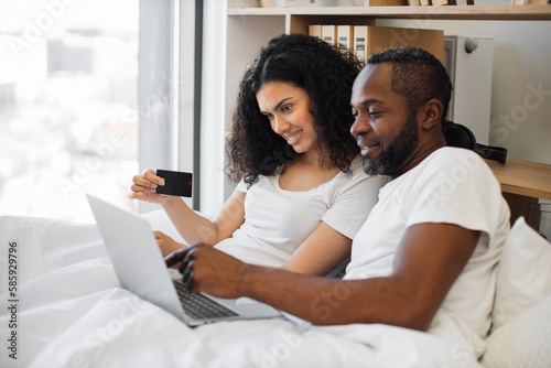 Loving multiethnic family spending morning in bed with laptop while using credit card for internet banking at home. Romantic partners making online payment operations in available application.
