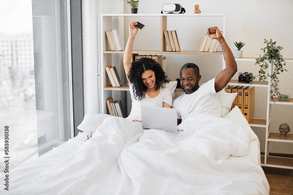 Emotional african american family of two raising hands in excitement while sitting in bed with laptop on blanket and credit card in hand. Thrilled married couple making successful deal online at home. Emotional african american family of two raising hands in excitement while sitting in bed with laptop on blanket and credit card in hand. Thrilled married couple making successful deal online at home.
