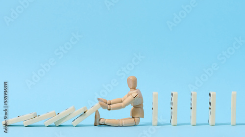 Fotografija A wooden puppet toy holds back a falling domino on a blue background, representi