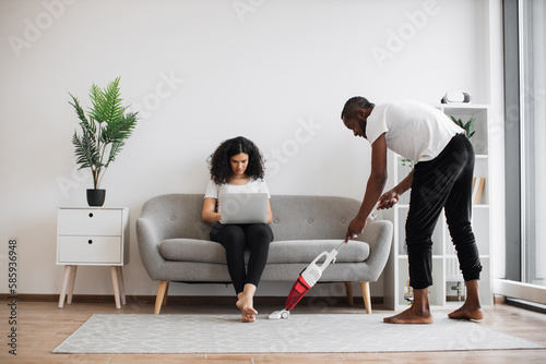 Side view of african man cleaning living room with cordless vacuum while focused woman working on laptop on couch. Caring husband doing households while wife freelancing at home.