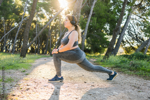 Adorable fat woman in tracksuit is engaged in fitness outdoor side view portrait copy space . Young overweight woman lunges outdoors on warm summer day. Healthy lifestyle and weight loss