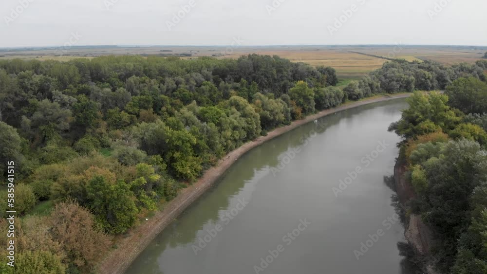 Plain river with banks and long narrow island overgrown with different trees at autumn sunny morning, aerial view when moving backwards.