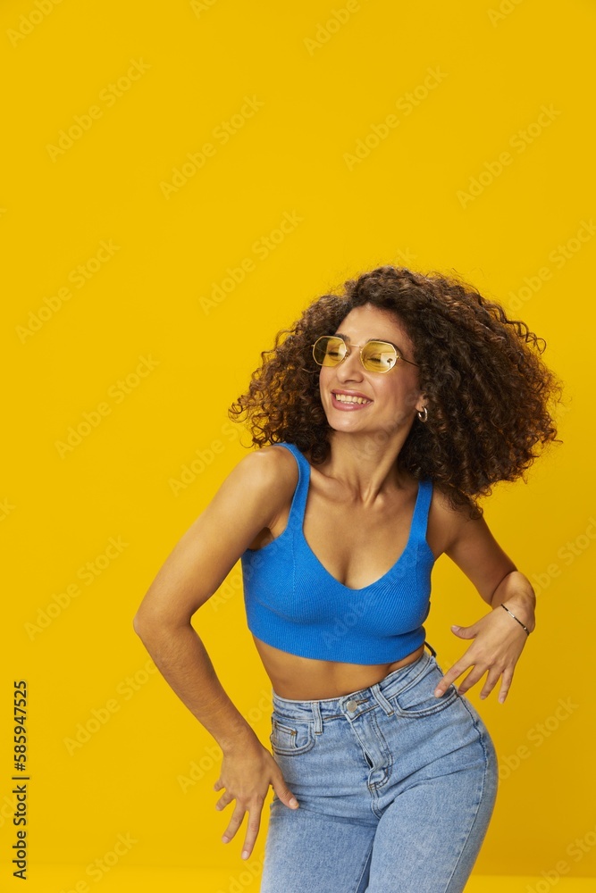 Woman with curly afro hair in a blue T-shirt on a yellow background dancing flying hair with sunglasses yellow, hand signs, look into the camera, smile with teeth and happiness, copy space Woman with curly afro hair in a blue T-shirt on a yellow background dancing flying hair with sunglasses yellow, hand signs, look into the camera, smile with teeth and happiness, copy space