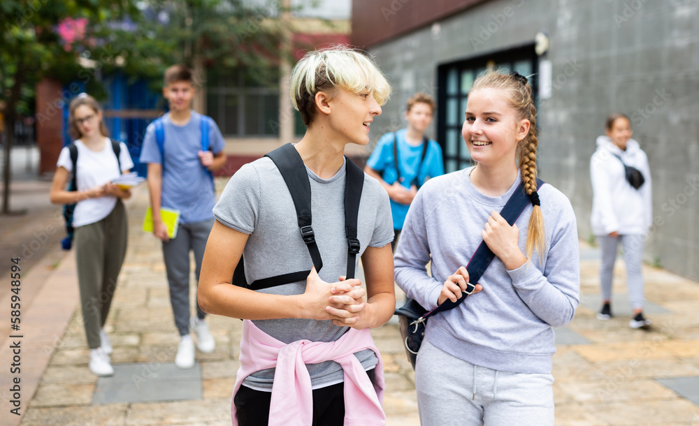 Portrait of teen female and male schoolmates with backpacks and workbooks walking to college campus on warm autumn day.