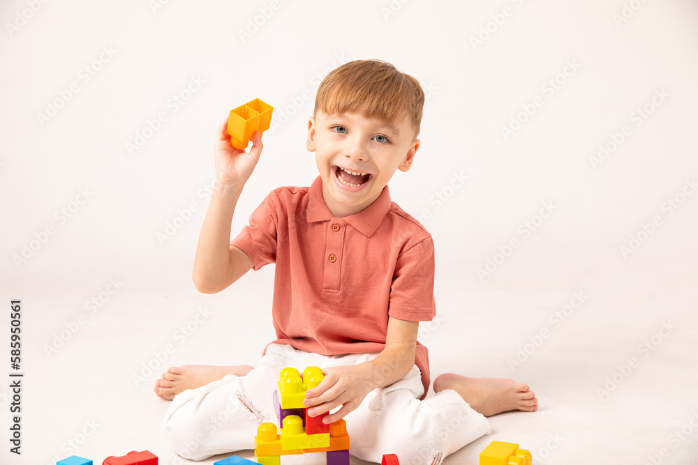 A happy child collects a tower of colored cubes. The preschooler sits down at the white background and assembles the construction set. The boy smiles, pleased with the result. A happy child collects a tower of colored cubes. The preschooler sits down at the white background and assembles the construction set. The boy smiles, pleased with the result.