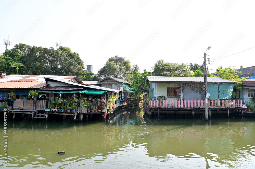 Obraz premium BANGKOK, THAILAND - MARCH 30, 2023 : Wooden houses, Thai houses and buildings along the Chao Phraya River with white clouds and blue sky background, Town Bangkok Waterfront, Thailand.