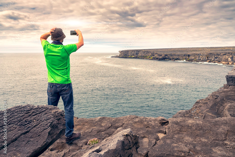 Male tourist in green shirt standing on edge of a cliff taking picture on his smart phone of stunning scenery. Aran Island, Ireland. Travel and tourism. Rough Irish nature landscape.