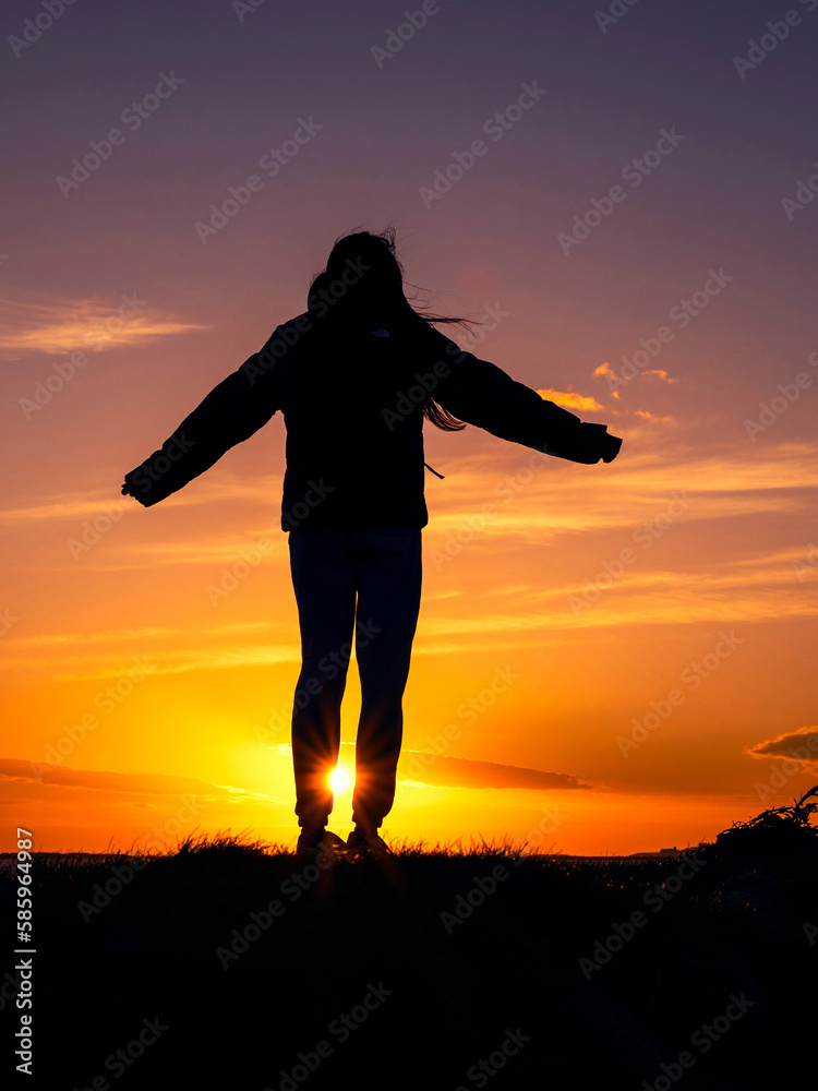 Silhouette of a teenager girl with long hair against sunset sky at a beach. Warm orange color. Enjoy outdoor concept. Beautiful nature scene at sunset. Low angle of view.