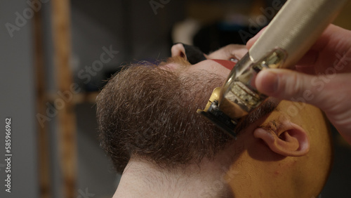 Professional barber trimming man's client beard in barbershop, close-up view. Cutting beard using hair clipper, electric trimmer machine. Hairdresser carefully cuts beard. Beauty hair salon for men.