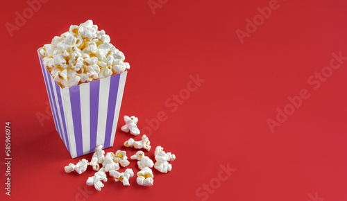 Purple white striped carton bucket with tasty cheese popcorn, isolated on red background. Box with scattering of popcorn grains. Fast food, movies, cinema and entertainment concept.