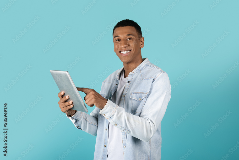 Cheerful millennial black guy in casual typing on tablet, surfing internet, chatting in social networks Cheerful millennial black guy in casual typing on tablet, surfing internet, chatting in social networks
