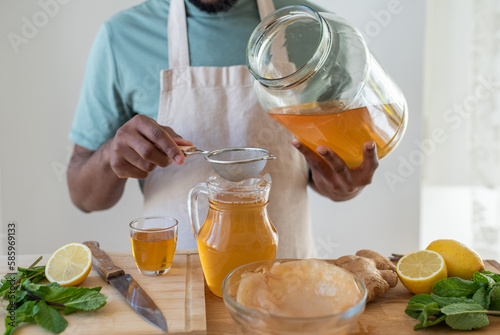 Making kombucha tea drink at home, fermented drink flavoured with ginger, lemon and mint. Healthy drink, organic probiotic beverage. Black young male hands pouring drink through a sieve.
